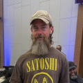 A man wearing a baseball cap displays a black t-shirt featuring the word "SATO$HI" above a Bitcoin symbol, visually representing the cryptocurrency theme referenced in the post "$XYZ those bags are heavy." The Bitcoin imagery reinforces the post's commentary about carrying heavy "bags" of crypto holdings.