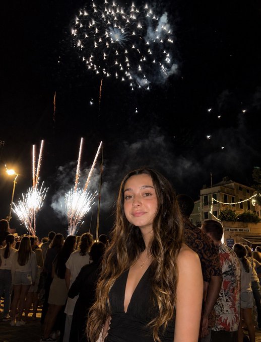 A young woman with long dark hair poses in the foreground while a fireworks display lights up the night sky above a crowd on a street.