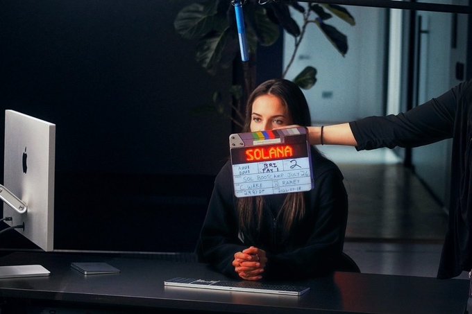 A clapperboard reading "SOLANA" is held up to a seated woman's face as she prepares for a video shoot, with the film slate marking "Sc 1, Tak 1" and date "2025-01-18." The image creatively depicts Solana as a subject being filmed, playing on themes of celebrity and attention in the crypto space.