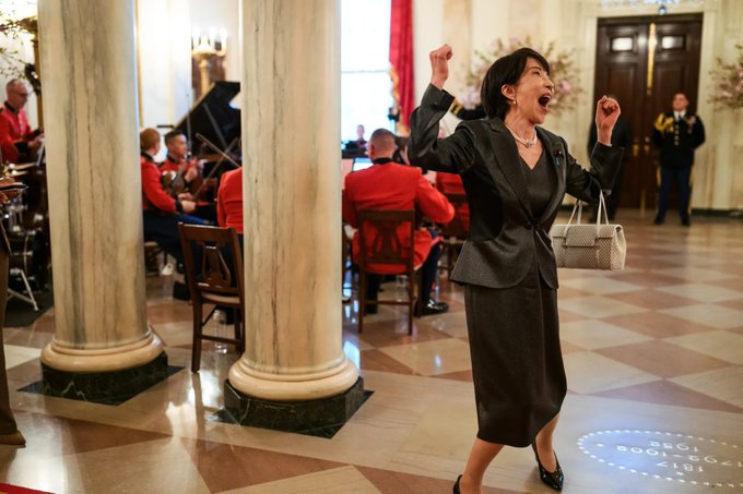 A woman in a black skirt suit throws her arms up with an ecstatic, open-mouthed expression in a grand marble-columned hall, as a military band plays in the background near a piano, while the social distancing sticker on the floor reads "1817-CROSSROOMS." The image captures a moment of over-the-top joy and disbelief, mirroring the post's rant about sleepless exhaustion and disbelief over Bitcoin ("that liar... sleeping 4-5 hours") tracking—you can almost imagine her erupting in triumph the moment BTC hits her target price.