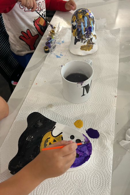 A close-up shows a hand using an orange paintbrush to mix purple paint on a palette, with a mug of dark liquid and a painted figurine visible on the paper-covered table.