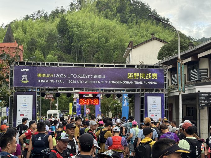 The image shows a large group of people gathered around an entrance gate, possibly at a race event or a popular tourist destination. The crowd consists of individuals wearing backpacks and hats, indicating that they might be participants or spectators. A sign can also be seen in the background, which reads "铜铃山トレングせずそちらします #STEPN #ランニング". The crowd appears to be waiting for something or someone, possibly a race start or an event organizer.