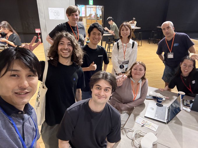 A group photo of about ten people posing together in what appears to be a coworking or event space, with some wearing lanyards and smiling at the camera.