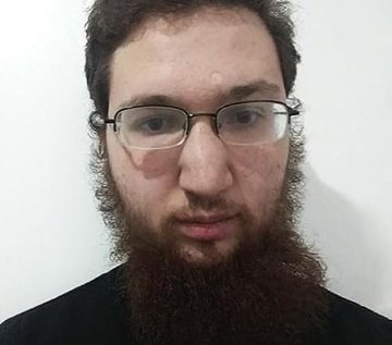 The image shows a man with glasses wearing a beard, standing against a white background. The man appears to be wearing a black shirt. There are no texts or other objects visible in the image.