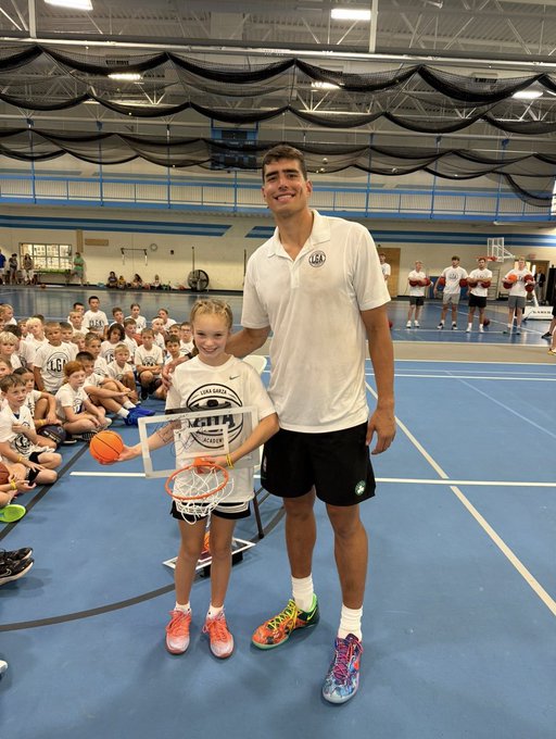 A young female camper and a tall male coach smile together at the "Fayette Camp" held in a gymnasium, with the girl holding a basketball and a small hoop while her shirt displays the camp's logo. The image captures a proud moment between mentor and participant, embodying the post's message about growth, loyalty to becoming a better player, and leaving past doubts behind.