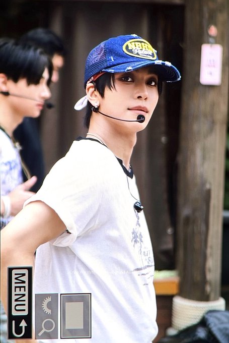 A young man wearing a blue trucker cap with a yellow "MTN" patch and a white t-shirt looks toward the camera while wearing a headset microphone. The cap's "MTN" branding and the post's hashtags "#XINLONG #鑫隆 #씬롱 #알파드라이브원 #ALPHADRIVEONE #ALD1 #알디원" suggest this is a member of the K-pop group ALD1, with the caption playfully referencing his handsome appearance.