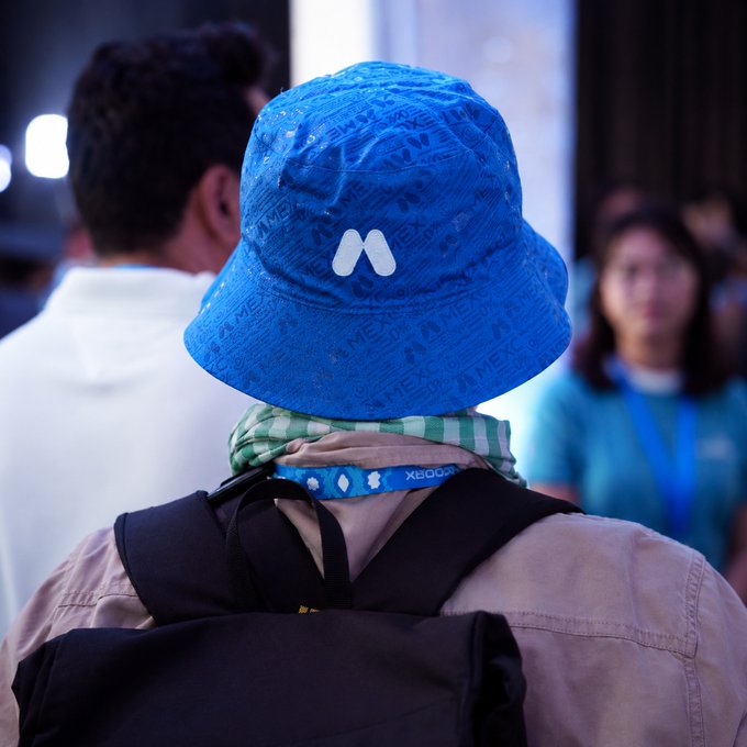 A person from behind wears a bright blue bucket hat featuring a repeating "MEXC" logo pattern and the exchange's white "M" emblem, paired with a green-and-white checkered scarf — clearly identifying them as a MEXCer. The promotional post perfectly matches the image, as the branded hat itself serves as the unmistakable giveaway of a MEXC community member.