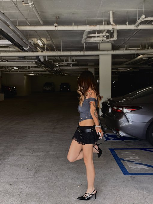 A woman in a gray top and black skirt poses in a parking garage next to a gray sports car with a handicap-accessible parking space nearby.