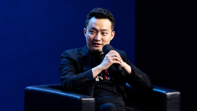 A man in a dark suit sits in a black armchair holding a microphone, speaking at an event against a blue backdrop.
