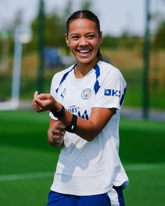 A Manchester City player wearing a white training jersey with Puma and OKX sponsor logos smiles brightly on a sunlit training pitch, adjusting her wristbands and fitness tracker. The image ties into the caption's message about daily training dedication and OKX's partnership, capturing an upbeat moment of preparation on the "green turf."