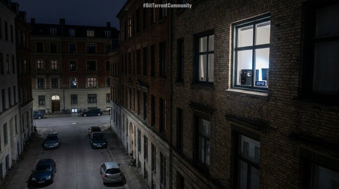 A nighttime view looking down at a courtyard surrounded by brick buildings, with cars parked below and one illuminated window showing a studio setup, marked '#BitTorrentCommunity'.