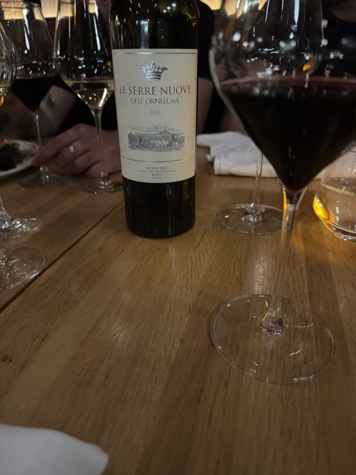A bottle of 2022 'Le Serre Nuove dell'Ornellaia' Bolgheri wine sits on a wooden table next to a glass filled with red wine.