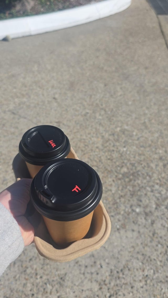 A first-person view of a hand carrying two takeaway coffee cups with black lids in a cardboard tray while walking along a paved path.