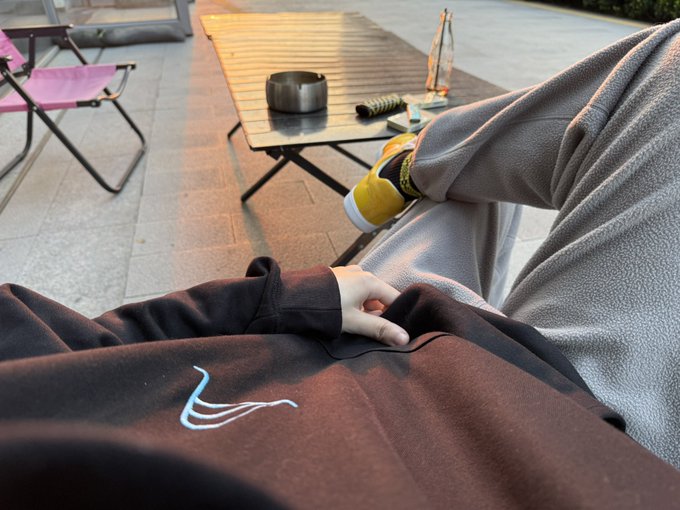 A casual POV shot of someone relaxing in a folding chair outdoors with a camping table holding a pot and thermos in the background, wearing yellow slippers and dark clothing.