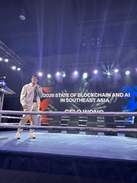 The image features an event where a man stands at a podium, giving a speech or presentation. The stage has a boxing ring setup behind him, indicating that this could be a boxing match or a similar sporting event taking place during his talk. The backdrop of the image is a large screen displaying information about blockchain and AI in Southeast Asia.
