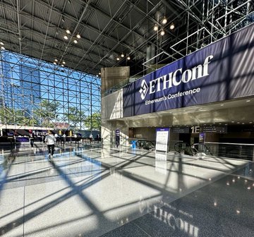 The image shows a large, open building with a glass ceiling that allows natural light to flood the space. A blue banner hangs prominently above the entrance, displaying the name "ETHGlobal" along with the words "EuthConference". The floor is made of concrete and reflects the sunlight, creating a bright atmosphere in the room.

There are several people walking around inside the building, some carrying backpacks or handbags. A few individuals can be seen near the entrance, while others are scattered throughout the space. In addition to these people, there are two bicycles parked within the building. The overall scene conveys a sense of activity and engagement with the conference happening in this large, open room.