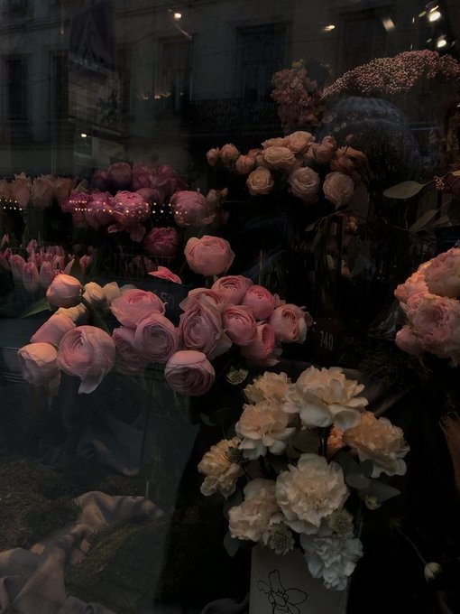 A display of flowers, including pink ranunculus, white roses, and peonies, is arranged in a shop window at night with reflections visible on the glass.