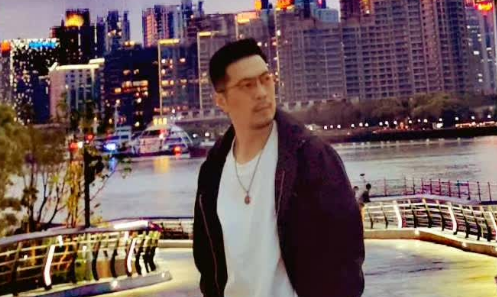 A man in a black jacket, white shirt, and glasses stands on a waterfront promenade at night with a brightly lit city skyline behind him.