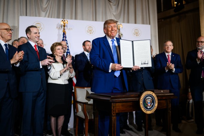 President Donald Trump holds up a signed document at a podium bearing the Presidential Seal, surrounded by clapping officials including Mike Johnson and Robert F. Kennedy Jr.