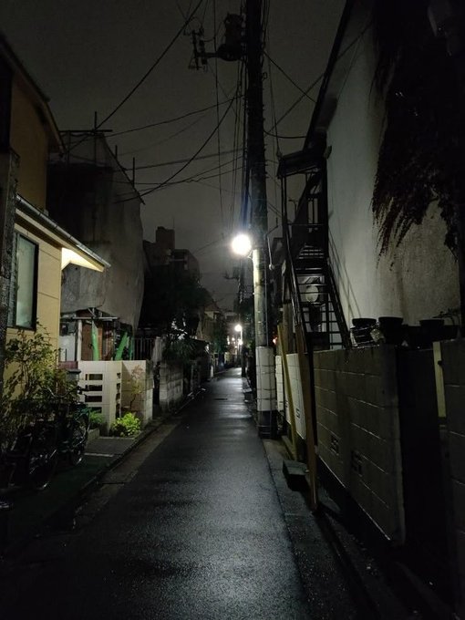 A narrow, dimly lit alleyway at night, illuminated by a single street lamp, with power lines crisscrossing overhead between residential buildings.