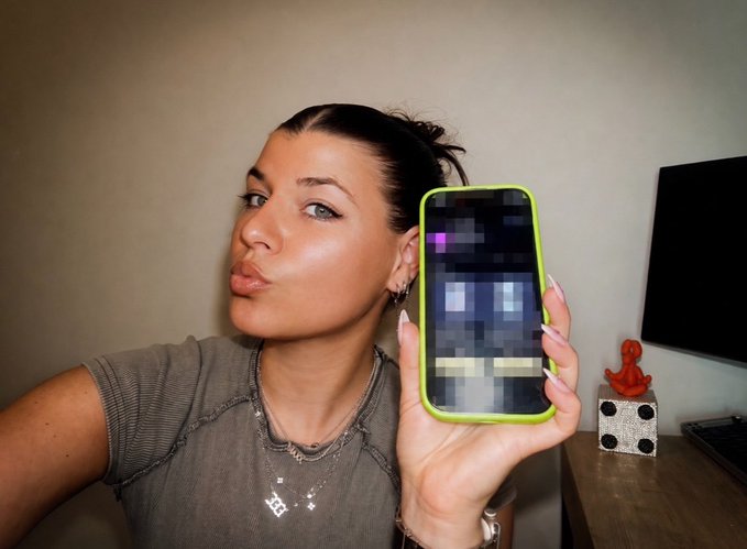 A woman takes a selfie while holding up a phone with a neon green case displaying a heavily pixelated image.