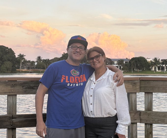 A smiling couple poses arm-in-arm on a wooden dock at sunset, the man wearing a blue 'Florida Gators' t-shirt and cap, the woman in a white blouse and glasses.