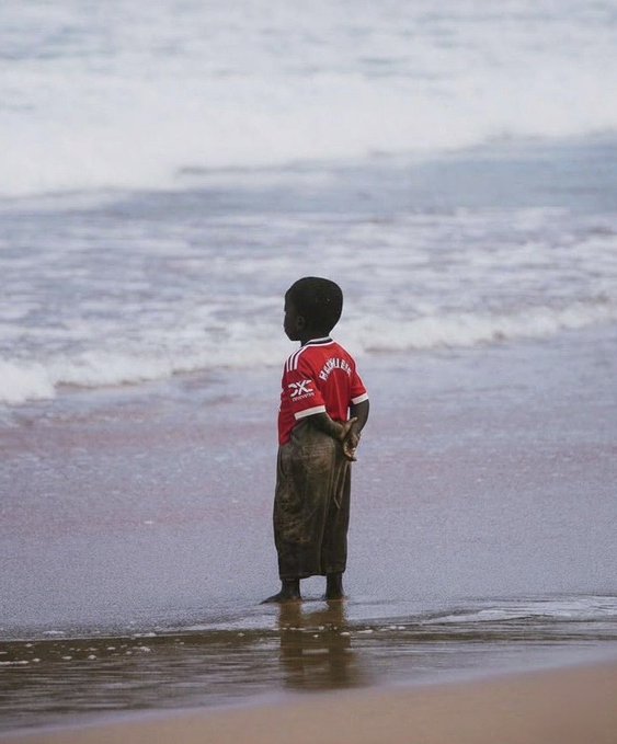 A young boy in a red "SK" jersey and dark shorts stands barefoot at the water's edge on a sandy beach, gazing out at the rolling waves. The serene, contemplative scene of a child watching the ocean evokes themes of patience and anticipation, subtly mirroring the post's message about Nomisma Network's evolution as the testnet countdown enters its final seven days.