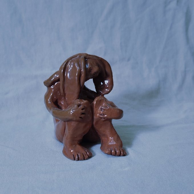A small brown clay sculpture of a hunched figure with their head resting on their knees, sitting on a light blue fabric surface.