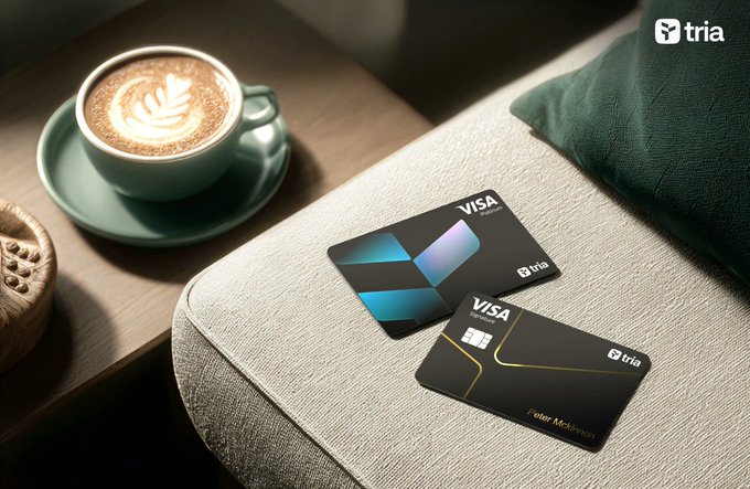Two black Visa credit cards from the brand 'tria' are placed on a light-colored cushion next to a green coffee cup and a pastry.