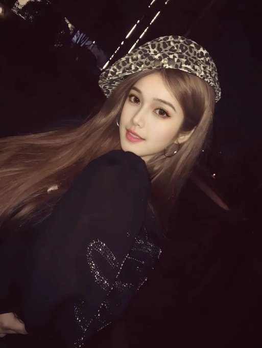 A selfie-style photograph features a young woman with long brown hair wearing a leopard-print hat and a black top, posing in a dimly lit setting.
