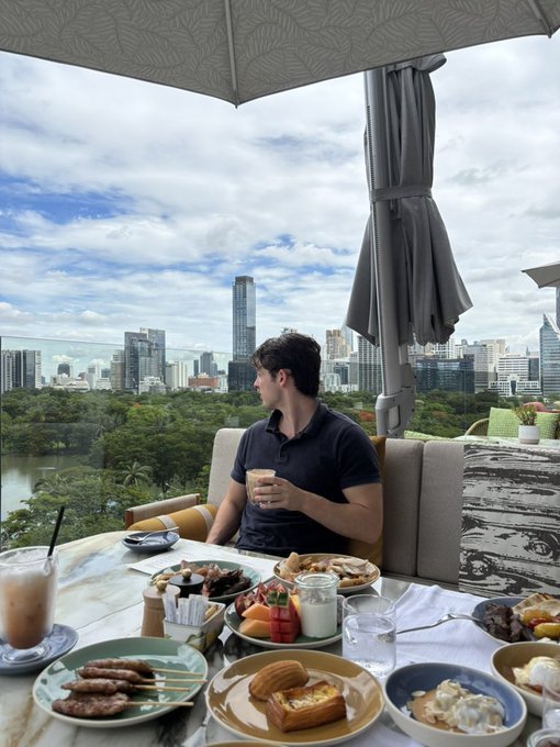 A young man in a dark polo shirt sits at a rooftop dining table filled with various breakfast dishes, holding a cup of coffee while gazing out at a city skyline featuring a lake and green park.