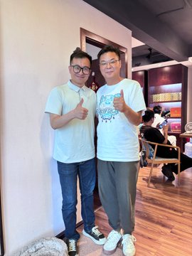The image features two men posing for a photo, both wearing white t-shirts with thumbs up signs. They are standing next to each other against a wall, which has a cat sitting on top of it. The background includes more people present but not as prominent in the foreground.