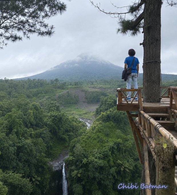 In this image, a person stands on a wooden deck overlooking a beautiful landscape with a waterfall. The person appears to be taking a moment to enjoy the view or possibly contemplating something. There are also other people present in the scene, but they seem to be further away from the main subject of interest.