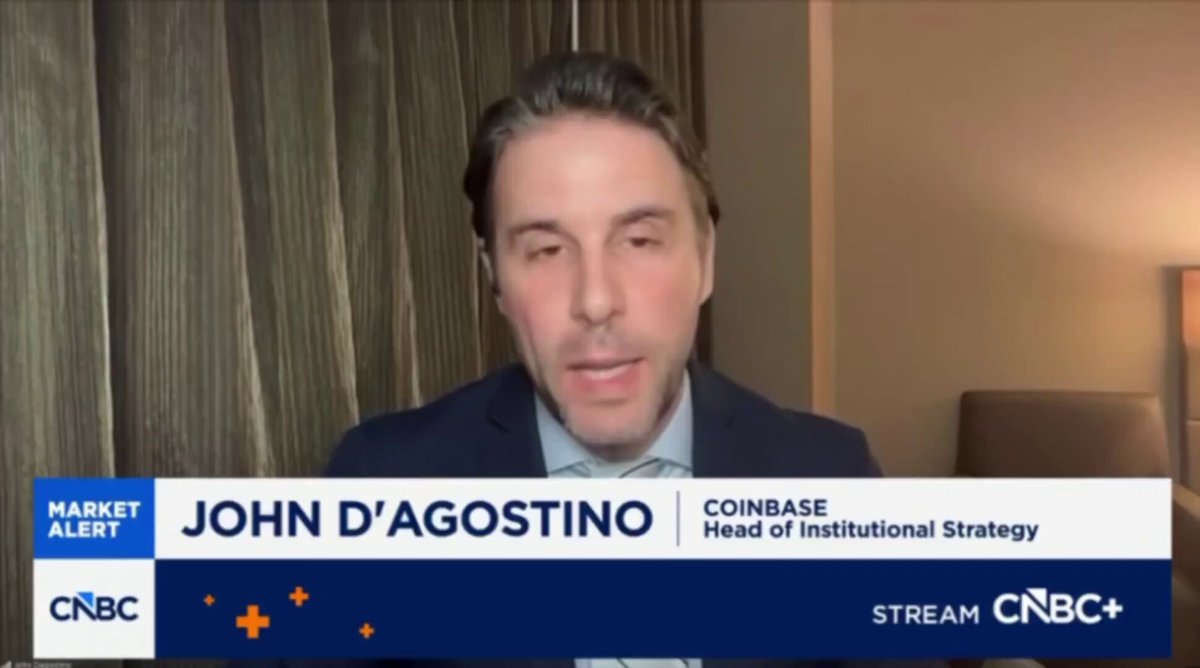 The image shows a man sitting behind a desk with a computer screen, talking into a microphone during an interview or webinar. The background features a brown curtain that partially covers part of the wall behind him. The main focus of the image is on John D'Agostino, who is speaking about coinbase and its appreciation in value over time.