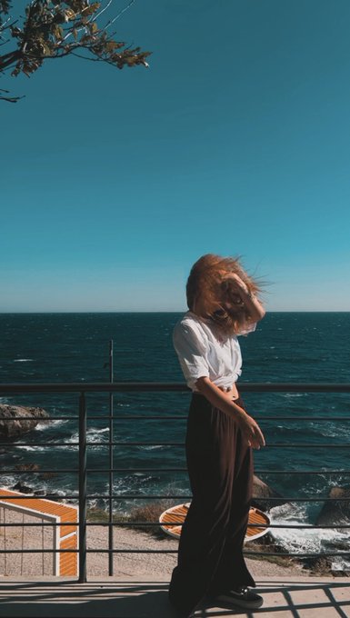 A person with windswept brown hair stands at a railing looking out at a deep blue ocean under a clear sky, wearing a white shirt and dark wide-leg pants.