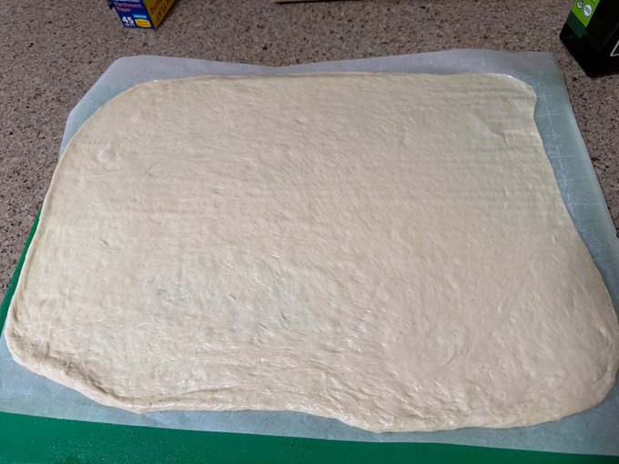 A photo of a rectangular sheet of dough rolled out flat on parchment paper over a green cutting board, with boxes visible in the background.