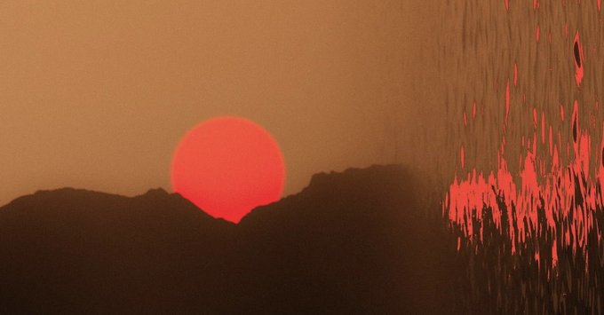 A red sun sets behind silhouetted mountains, mirrored in rippling water on the right side of the frame. The hazy, atmospheric scene evokes a quiet market close—fitting for a Twitch stream focused on watching charts and chatting about trading.