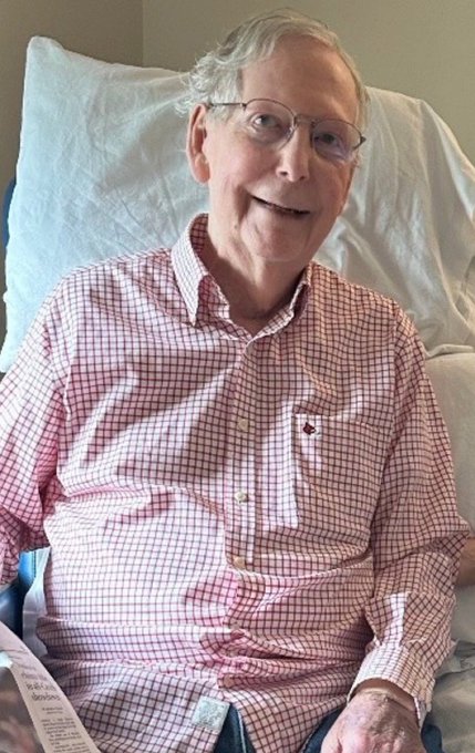 A photograph of an elderly man with white hair and glasses, smiling while reclining in a hospital bed, wearing a pink and white checkered button-up shirt.