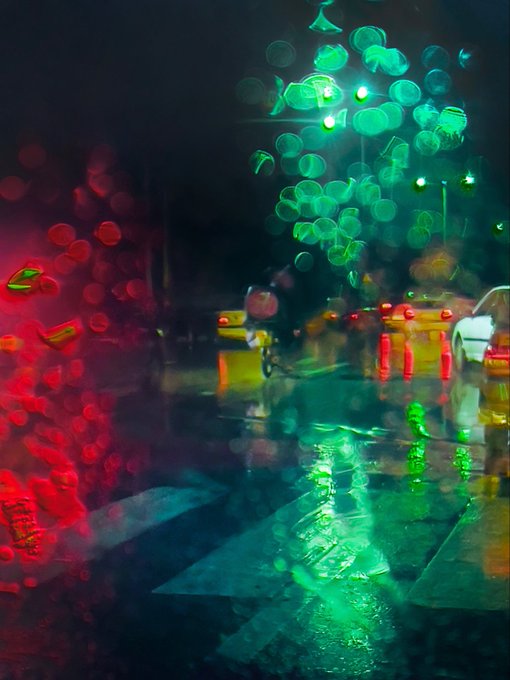 A blurry, atmospheric photo taken through a rain-covered window at night, showing out-of-focus red and green traffic lights reflecting on a wet street.