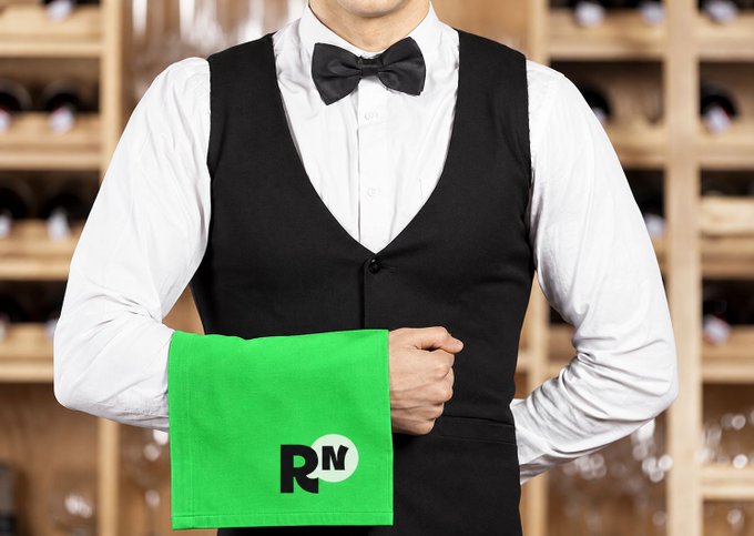 A waiter in a white shirt, black vest, and bow tie holds a bright green napkin featuring a 'RN' logo, standing in front of a wine rack.