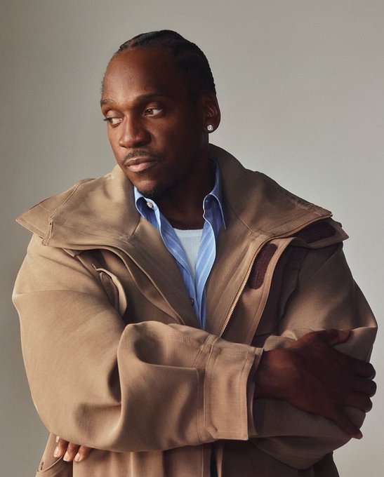 A portrait of a man with short hair wearing a tan jacket over a blue striped shirt, standing with his arms crossed against a neutral background.