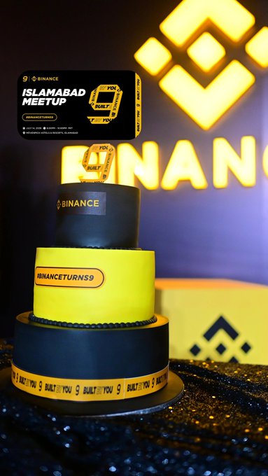 A custom Binance-branded tiered cake in black and yellow celebrates Binance's 9th anniversary at the Islamabad Meetup, featuring "BINANCETURNS9," "ISLAMABAD MEETUP," and "BUILD" text alongside the iconic Binance logo, commemorating the company's milestone with the local crypto community.