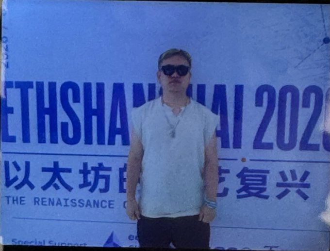 A man wearing sunglasses and a white polo shirt stands in front of a blue backdrop displaying text including 'ETHSHANGHAI 2023' and 'THE RENAISSANCE'.