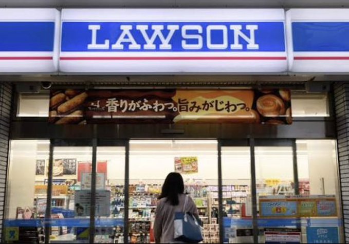 A customer enters a Lawson convenience store with the iconic blue "LAWSON" signage prominently displayed above the entrance, alongside a Japanese promotional banner reading "香りがふわっ、旨みがじわっ" (fragrant aroma, rich flavor) for in-store baked goods. The image visually represents the Tokyo retail location where Lawson is piloting JPYC stablecoin payments in collaboration with KDDI and HashPort.