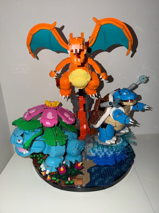 A LEGO display features three iconic Pokémon — Charizard with outstretched orange and teal wings, Venusaur with a pink flower, and Blastoise with water cannons — posed on a detailed base with grass and water elements, perfectly capturing the excitement of shouting "MINA MINA MINAAAAAAAAAAA" as if summoning these classic Kanto starter evolutions.