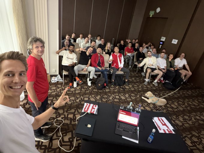 A man in a white t-shirt smiles and flashes a peace sign in the foreground, while a group of around 20 colleagues, some wearing red shirts, gather behind a table with laptops displaying "RedStone," a conference room number "04" placard, and a golden retriever wearing a blue bandana. The scene captures the team bonding at the RedStone offsite, reflecting the post's message about the team's high energy and excitement for what's ahead on Day 1.