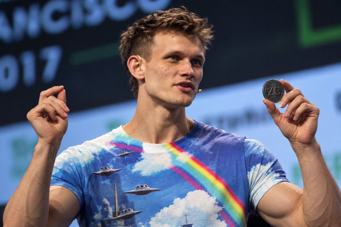 A young man with blonde hair wearing a blue t-shirt with a rainbow and UFO design holds a ZC (Zcash) coin at what appears to be a cryptocurrency conference.