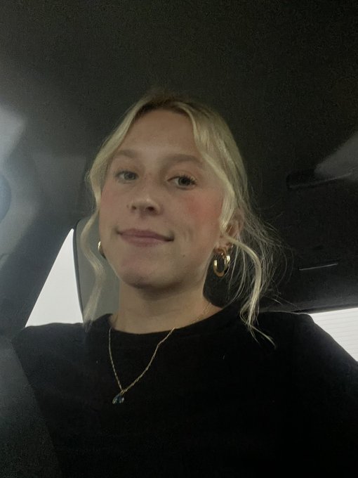 A young woman with blonde hair wearing a black top and gold jewelry takes a selfie inside a car.