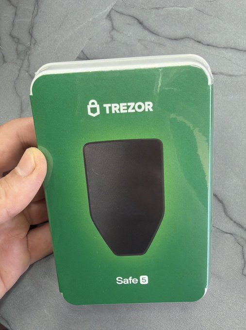 A hand holding a sealed green package for a Trezor Safe 5 hardware wallet.