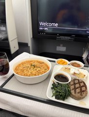 An airline meal of noodles, greens, and grilled chicken is served on a tray with a "Welcome" screen displayed in the background, juxtaposing the relaxing in-flight dining experience with the post's message about the low-energy approach of doing nothing in a trading competition hosted by @MEXC.